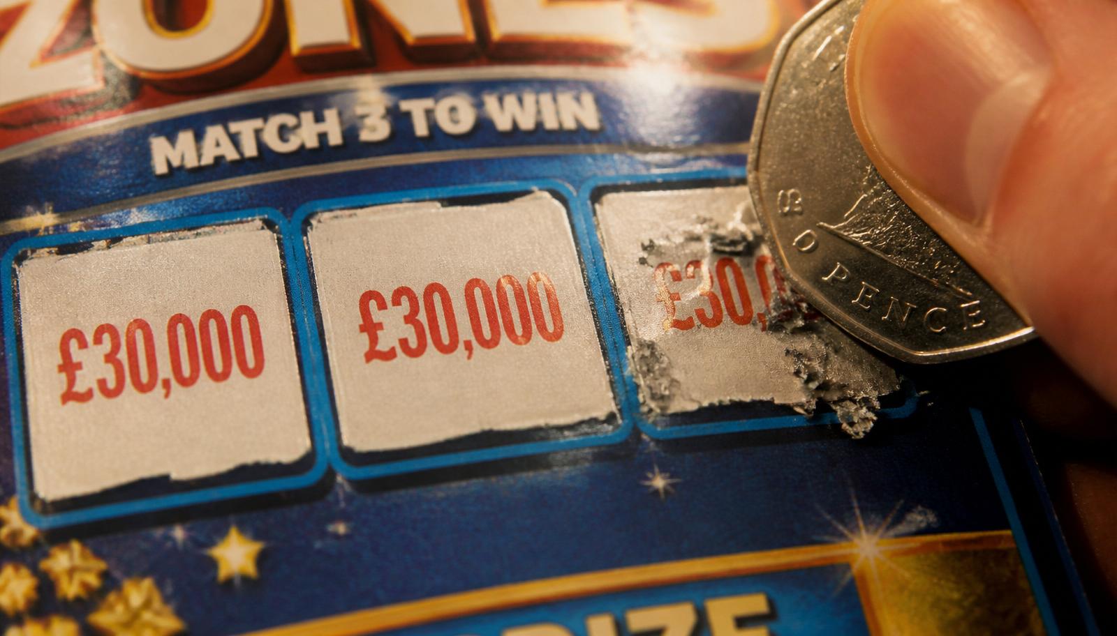 Macro close-up of a thumb and forefinger holding a 50p coin scratching a match-three lottery card — first two panels revealed as £30,000, third panel mid-scratch